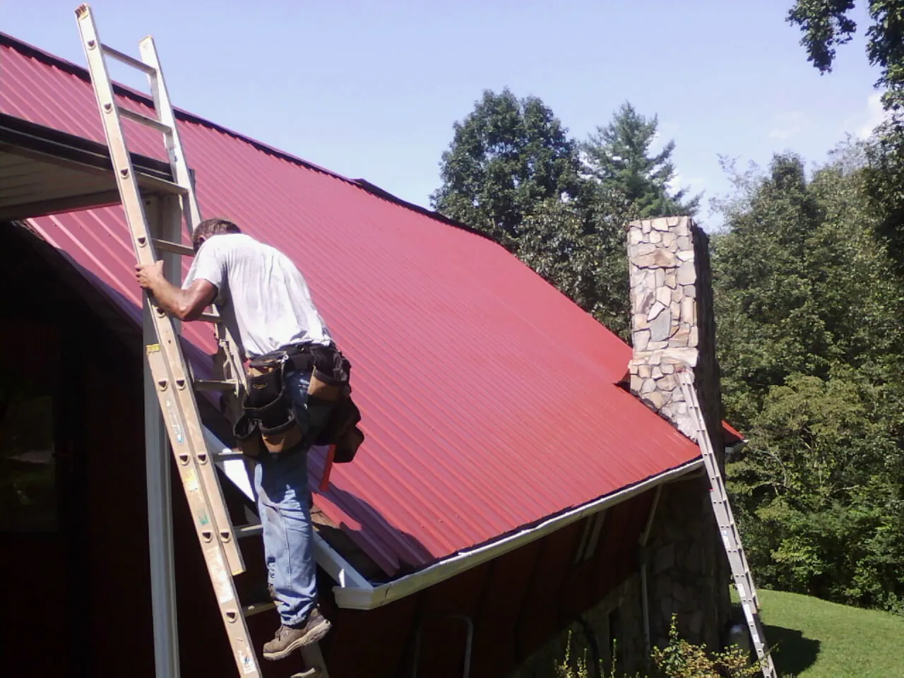 Roofer on a ladder beside a red metal roof and stone chimney — Soffit & Fascia Repair by Roofing Brothers in Ringgold
